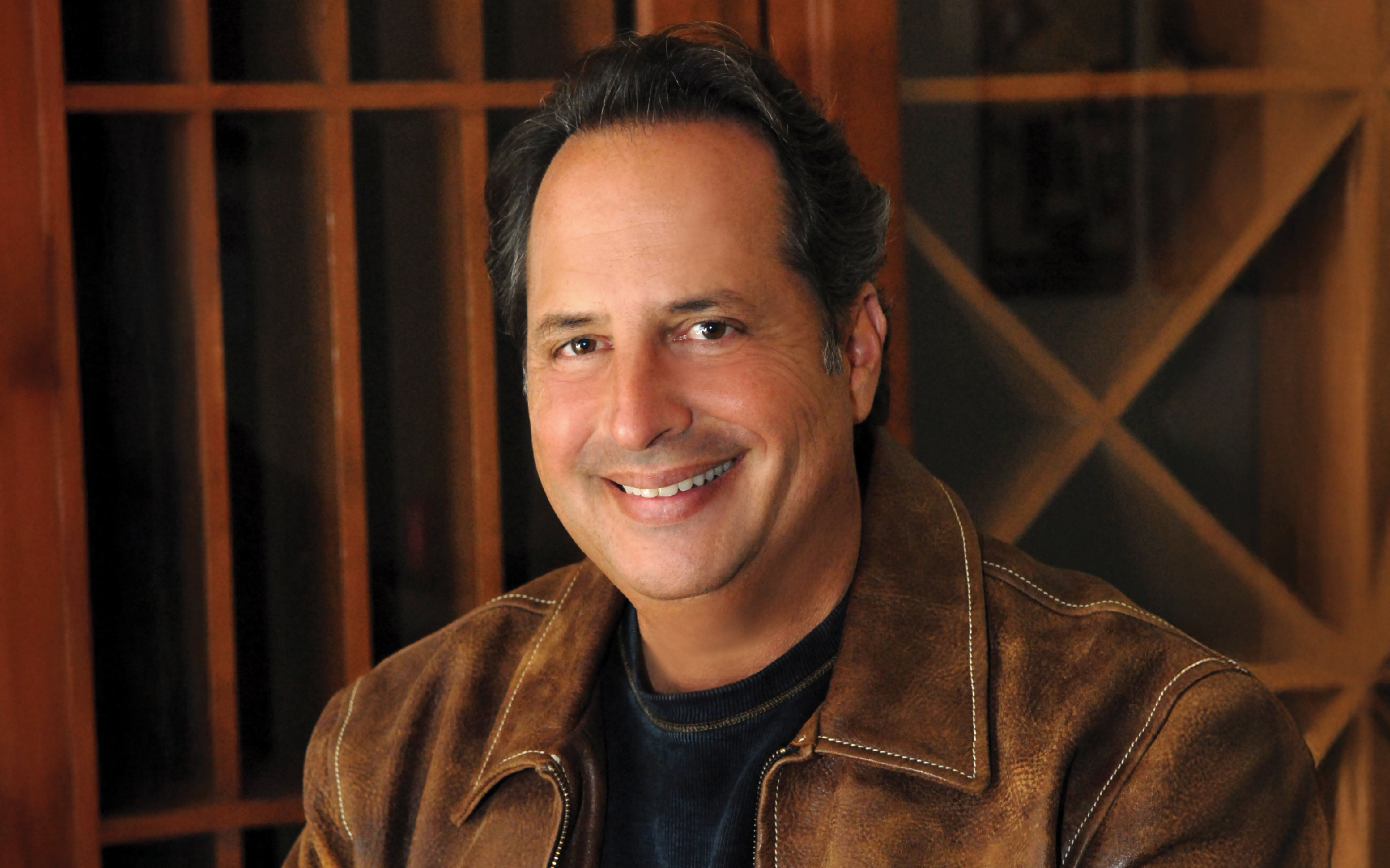 Comedian Jon Lovitz is photographed smiling, wearing a black shirt and brown leather jacket. The background includes wooden fixtures.