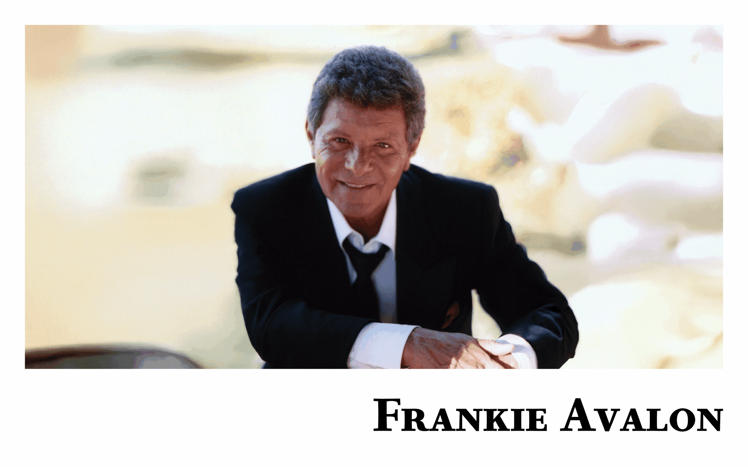 Singer Frankie Avalon, dressed in a black suit, white shirt, and loosened black tie, smiles warmly while leaning forward with one arm resting on a chair. The soft, blurred background gives a sunlit, golden glow. His name ‘Frankie Avalon’ is written in bold black letters at the bottom of the image.