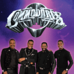 Four members of The Commodores stand smiling in front of a purple background with the band’s large metallic logo above them. They are dressed in coordinated black outfits with sparkling accents.